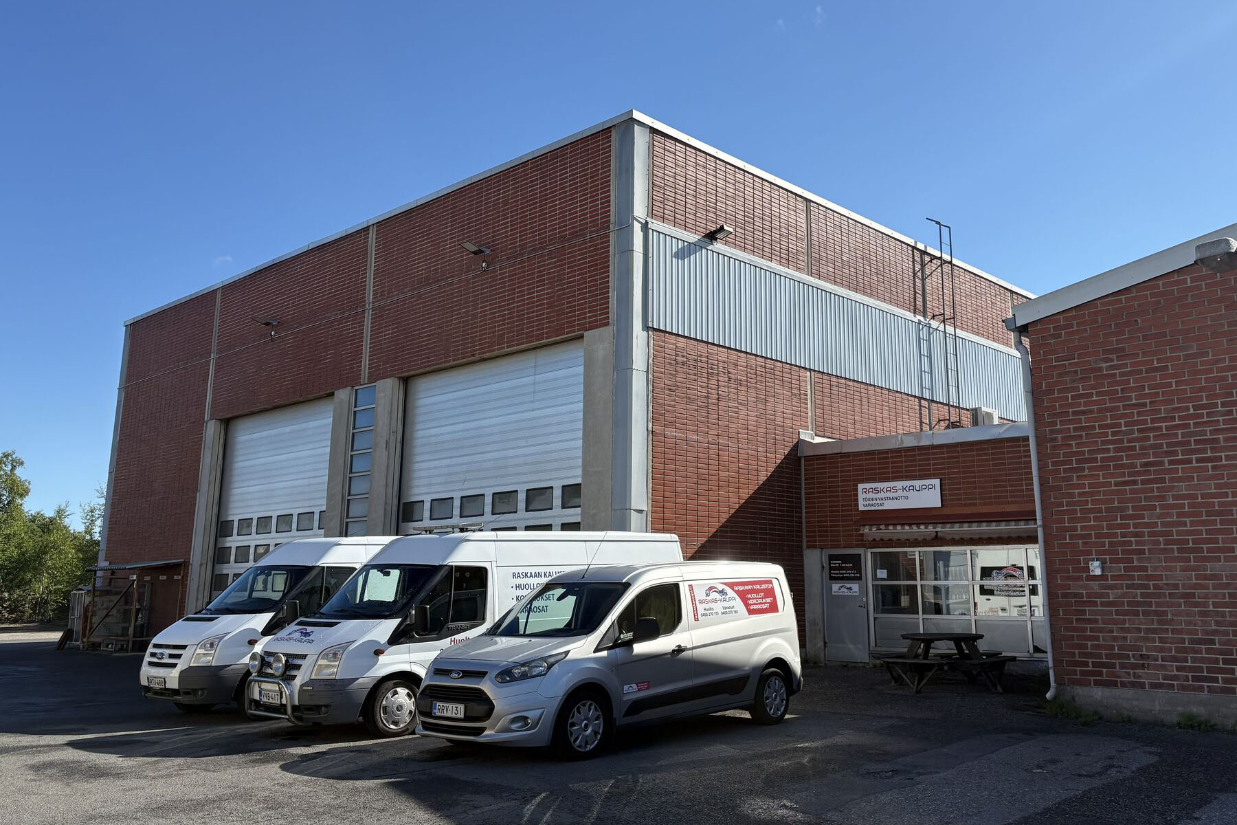 Brick industrial warehouse with large roll-up doors and three white delivery vans parked outside on a sunny day.